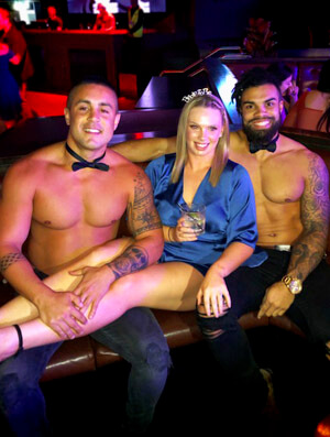 two topless waiters wearing bow ties with the hen spread across their laps in a darlinghurst nightclub